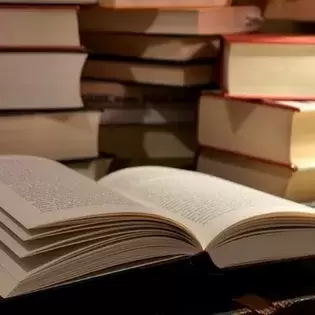 An open book on a table with several stacks of books behind it. 